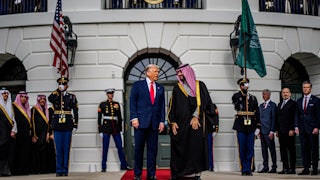 Donald Trump and Saudi crown prince Mohammed bin Salman stand on a red carpet in front of the White House. Members of the U.S. military and MBS's team stand in the background.