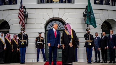 Donald Trump and Saudi crown prince Mohammed bin Salman stand on a red carpet in front of the White House. Members of the U.S. military and MBS's team stand in the background.