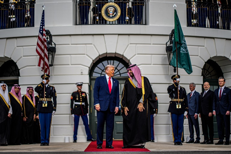 Donald Trump and Saudi crown prince Mohammed bin Salman stand on a red carpet in front of the White House. Members of the U.S. military and MBS's team stand in the background.