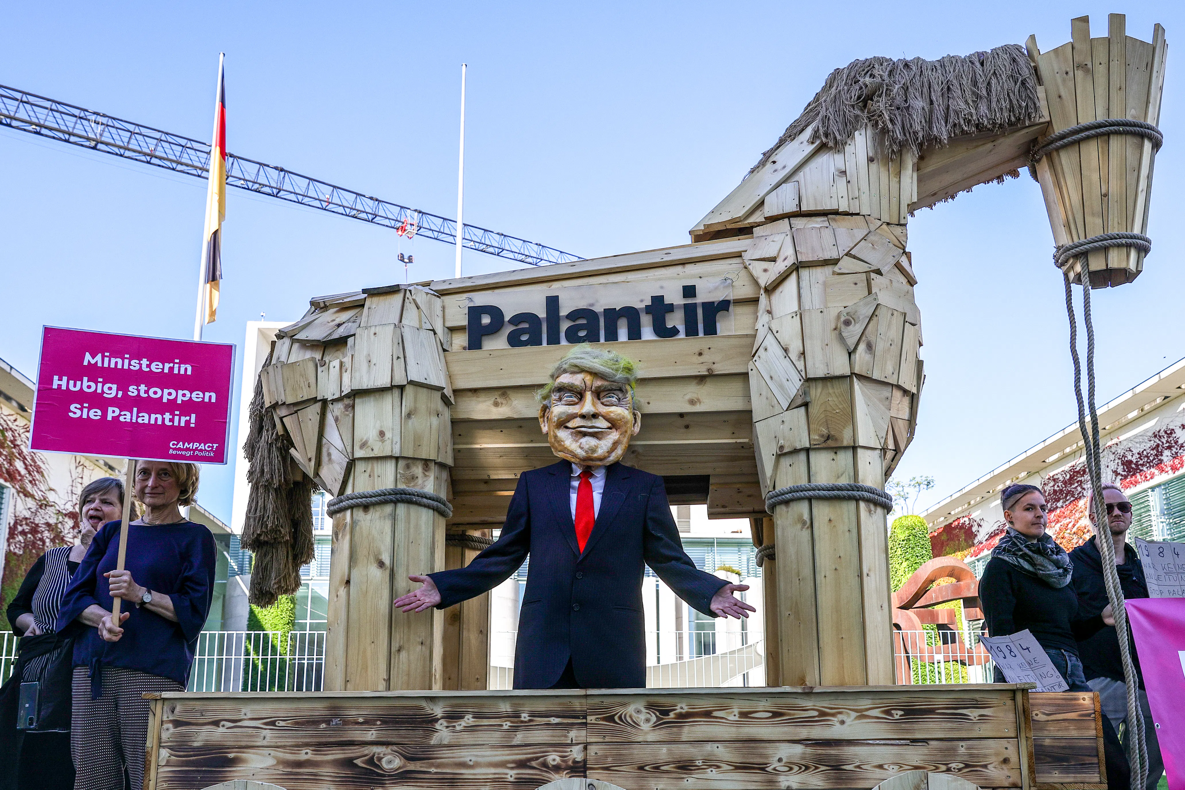 A man dressed as Donald Trump stands in front of a wooden horse with Palantir written on it at a protest in Berlin, Germany, Another protestor stands to the left holding a picket sign in German.