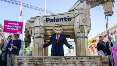 A man dressed as Donald Trump stands in front of a wooden horse with Palantir written on it at a protest in Berlin, Germany, Another protestor stands to the left holding a picket sign in German.