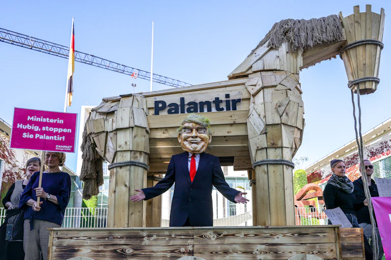 A man dressed as Donald Trump stands in front of a wooden horse with Palantir written on it at a protest in Berlin, Germany, Another protestor stands to the left holding a picket sign in German.