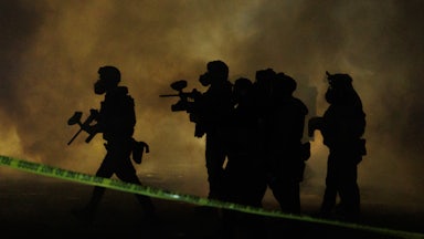 Six federal agents wearing gas masks walk through tear gas at night.