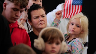 A white South African woman holds a small blonde child waving a U.S. flag. Other white South african children and adults stand near them.