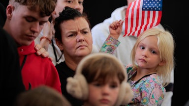 A white South African woman holds a small blonde child waving a U.S. flag. Other white South african children and adults stand near them.