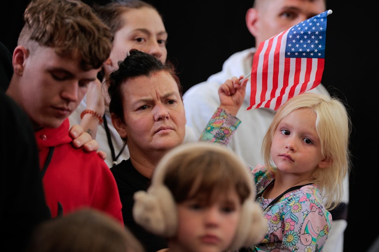 A white South African woman holds a small blonde child waving a U.S. flag. Other white South african children and adults stand near them.