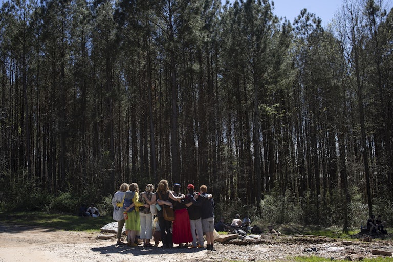 Demonstrators stand with their backs to the camera, their arms around one another, in front of trees.