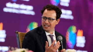 Amodei sits at a table with flowers and gestures while turning his head to his right and speaking into a microphone.