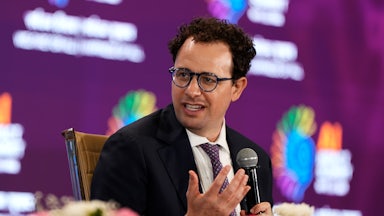 Amodei sits at a table with flowers and gestures while turning his head to his right and speaking into a microphone.