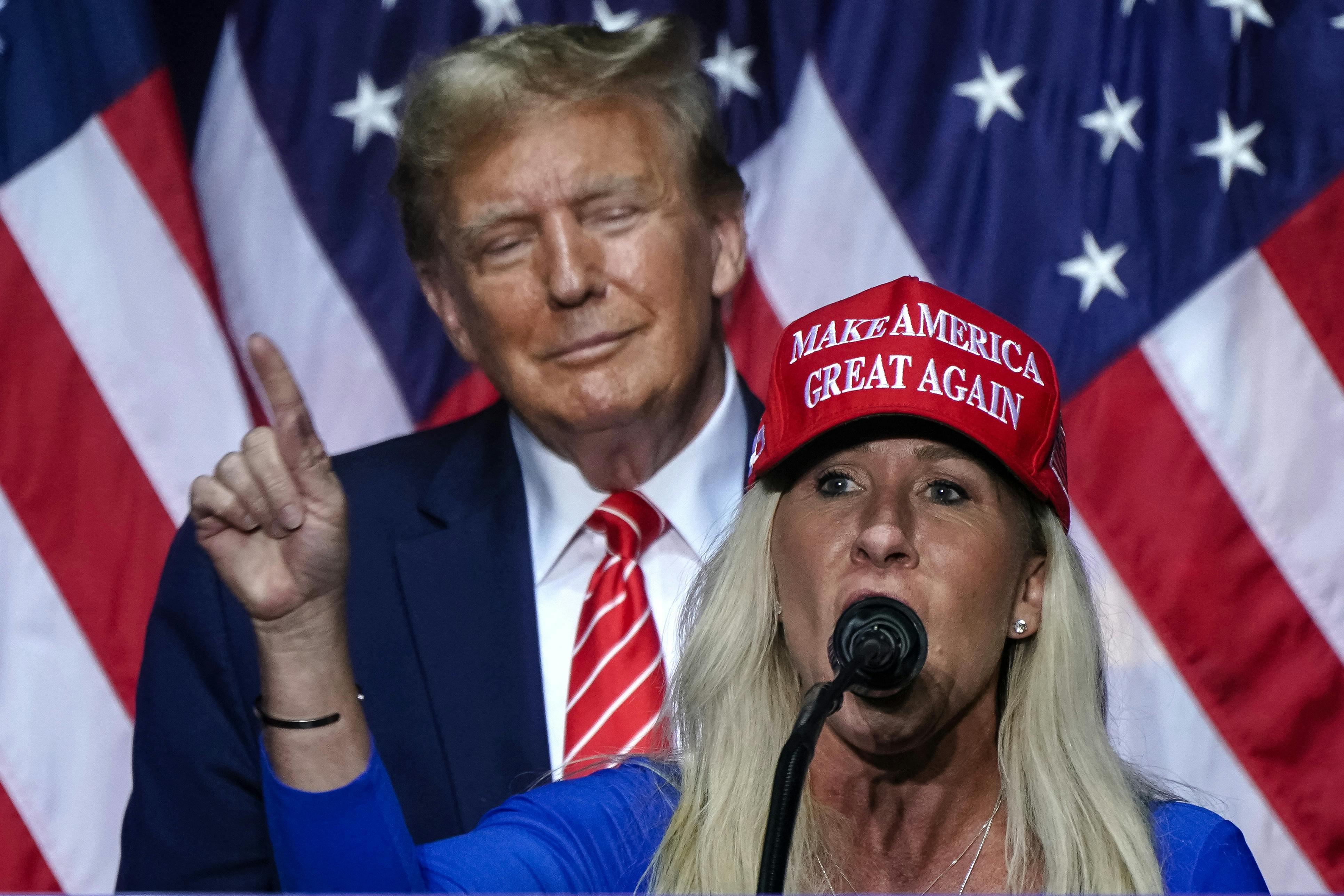Donald Trump smiles as he stands behind Marjorie Taylor Greene, who is giving a speech.