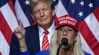 Donald Trump smiles as he stands behind Marjorie Taylor Greene, who is giving a speech.