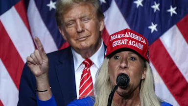Donald Trump smiles as he stands behind Marjorie Taylor Greene, who is giving a speech.