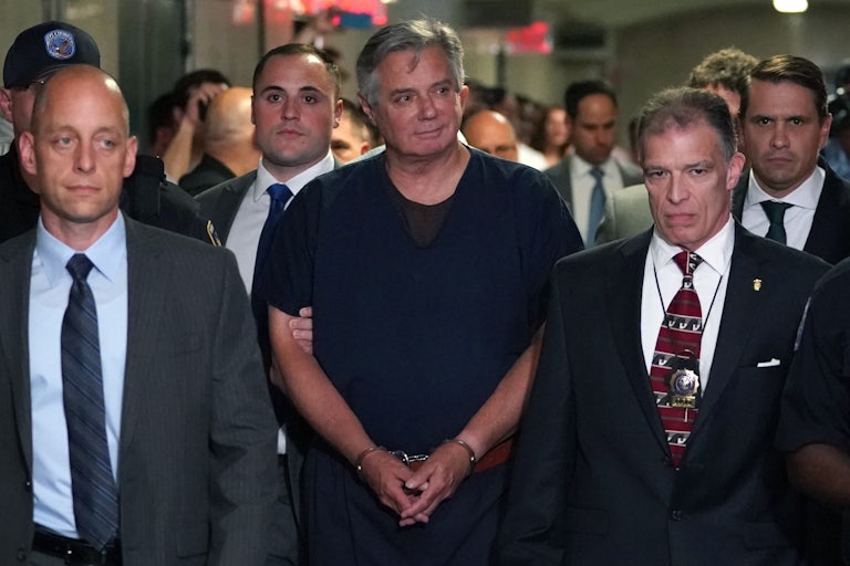 Paul Manafort walks in handcuffs and smiles at something off camera. Several others around him, including a security guard who holds his arm.