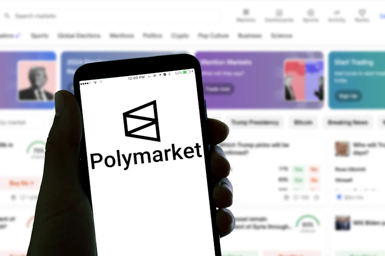 A hand holds a phone with the Polymarket logo. A screen in the background also shows the Polymarket homepage, featuring an image of Donald Trump.