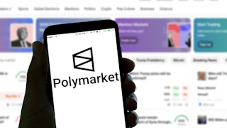 A hand holds a phone with the Polymarket logo. A screen in the background also shows the Polymarket homepage, featuring an image of Donald Trump.
