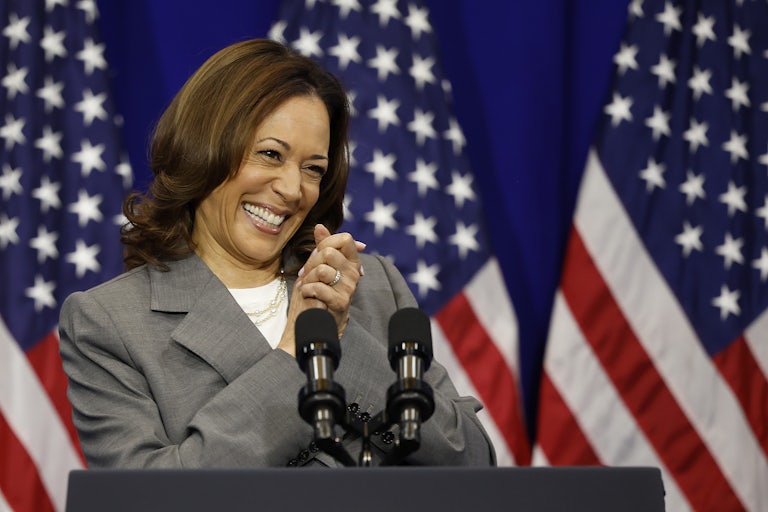 Kamala Harris clasps her hands and smiles in pure joy. A lectern is in front of her and three U.S. flags are behind her.