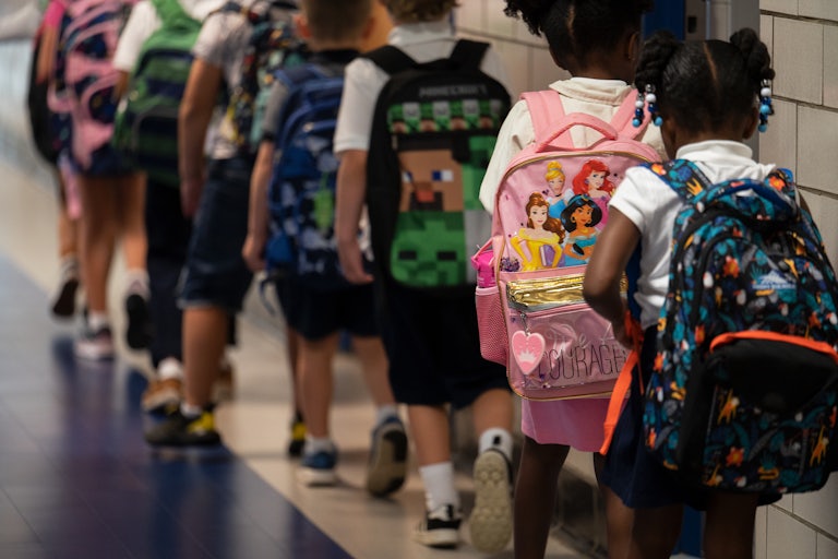 A group of young students (perhaps pre-school age) wear their backpacks and walk in a single-file line in school.
