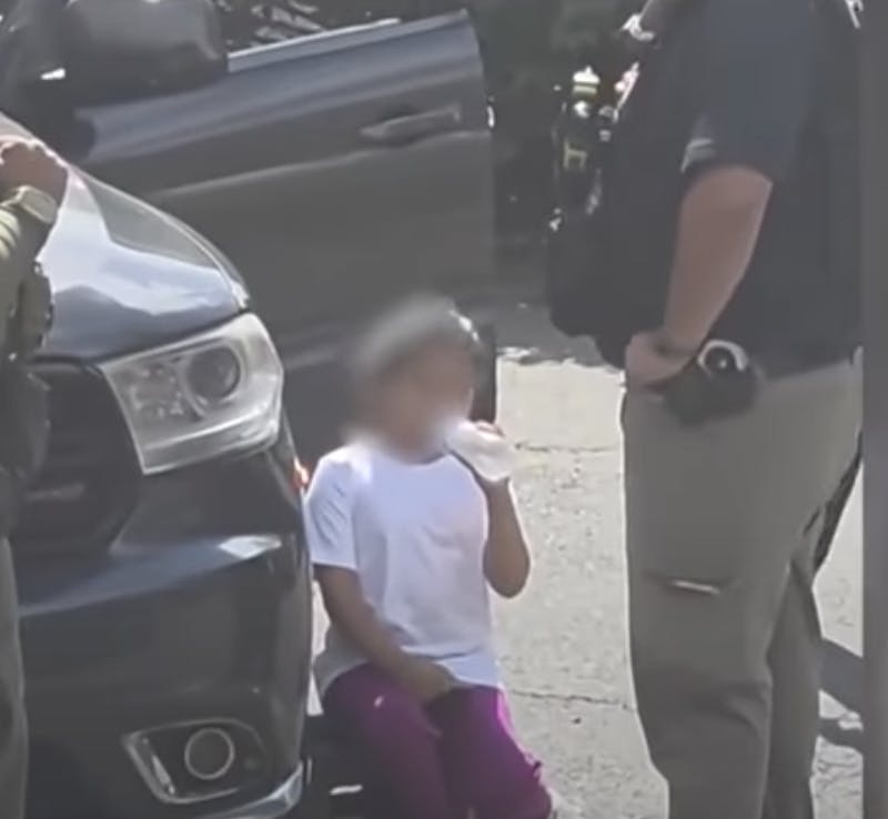 A small child with her face blurred out drinks milk out of a bottle while standing next to a car. Two other agents can be seen the video.