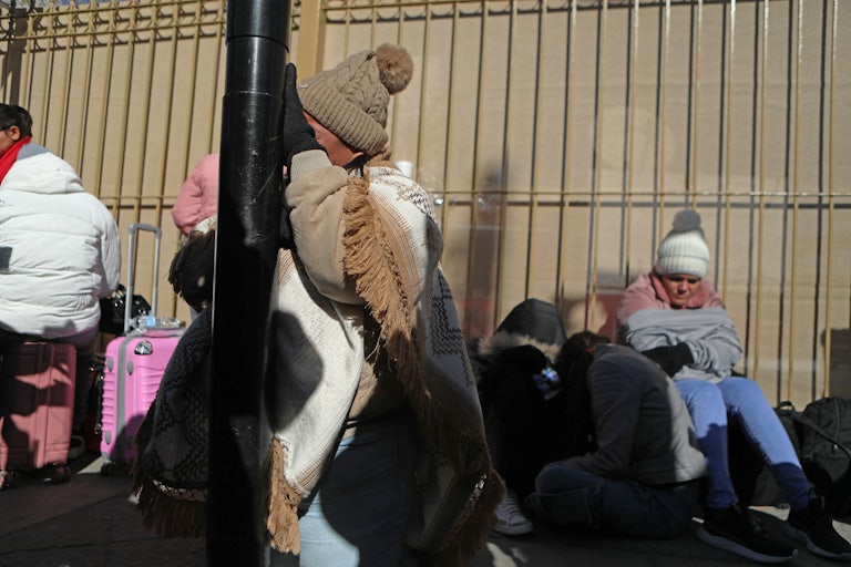 A woman cries as she leans against her head against a pole. Another woman sits in the background huddled by a fence.