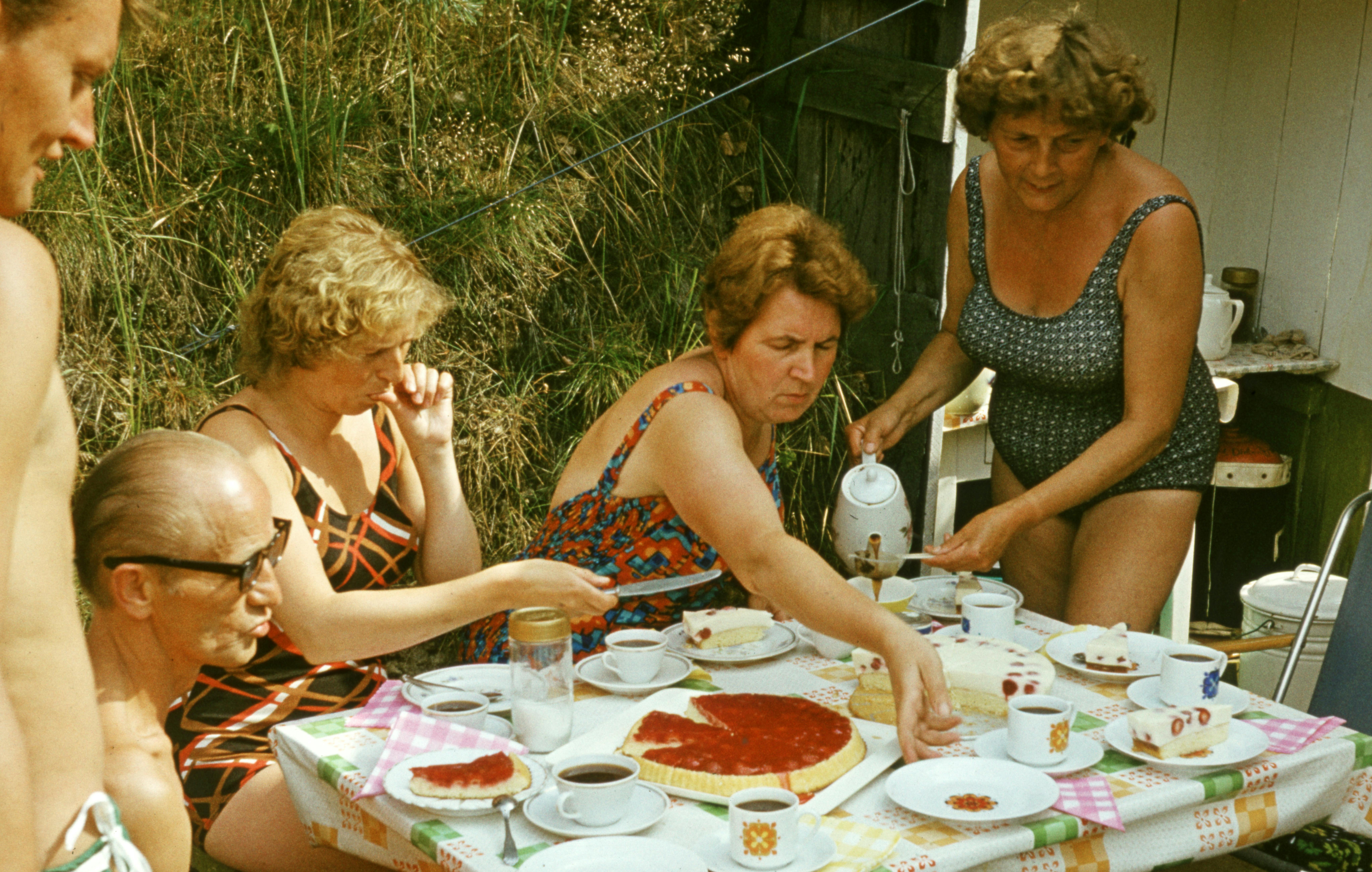 Five adults gather in swimsuits around a small table with cake on it.