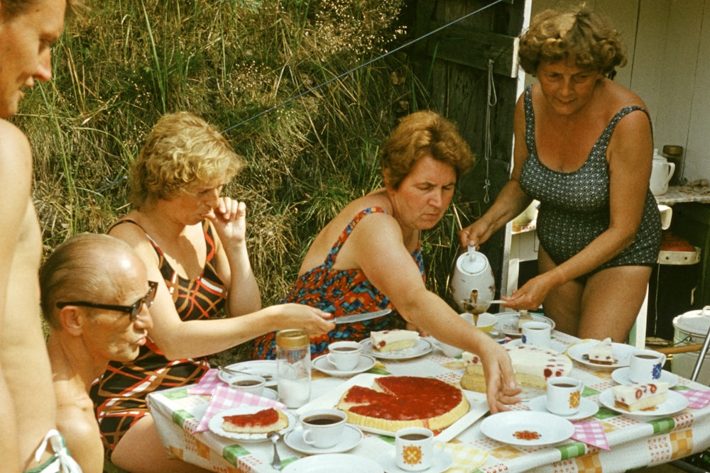 Five adults gather in swimsuits around a small table with cake on it.