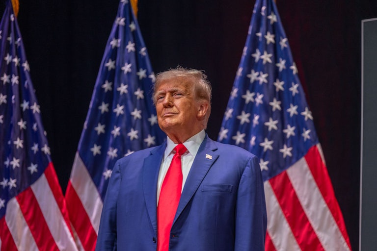 Donald Trump smiles proudly as he stands in front of a row of U.S. flags.