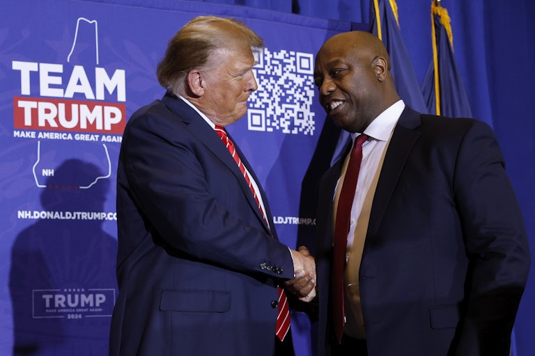 Donald Trump and Tim Scott shake hands on stage. In the background is a backdrop that says Team Trump with a QR code.