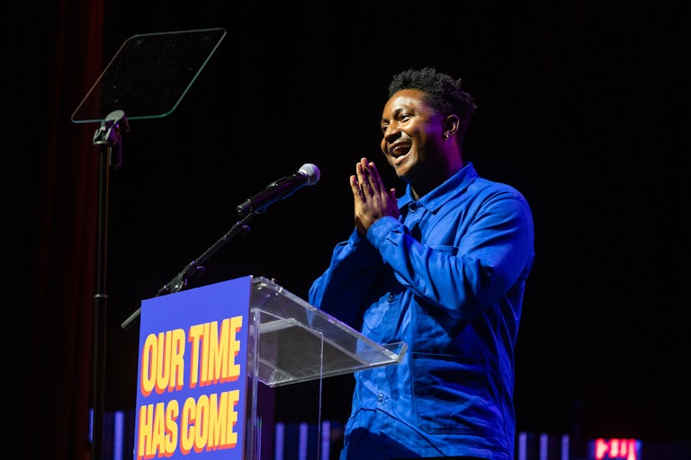 Chi Ossé smiles and puts his hand together in a prayer gesture at a podium that reads "Our Time Has Come."
