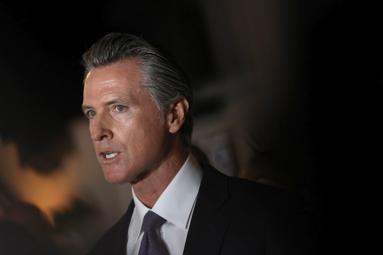 A close-up of Gavin Newsom standing in shadows as he speaks at a press conference.
