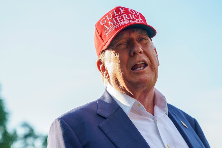 Donald Trump speaks outside of the White House while wearing a red cap that reads "Gulf of America | Yet Another Trump Development."
