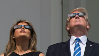 Donald Trump and Melania Trump stand side by side, both looking upwards while wearing protective eclipse glasses.