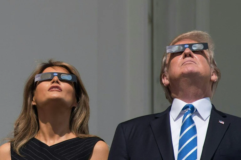Donald Trump and Melania Trump stand side by side, both looking upwards while wearing protective eclipse glasses.