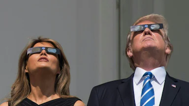 Donald Trump and Melania Trump stand side by side, both looking upwards while wearing protective eclipse glasses.