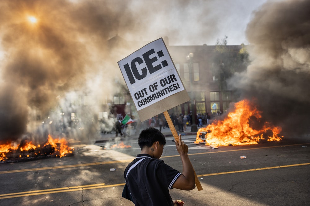 On June 8, a young man in downtown Los Angeles held a sign that reads “ICE: Out of Our Communities!” as smoke from burning Waymo driverless cars filled the air.