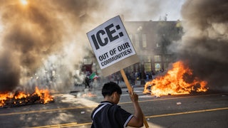 On June 8, a young man in downtown Los Angeles held a sign that reads “ICE: Out of Our Communities!” as smoke from burning Waymo driverless cars filled the air.