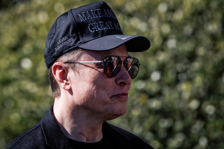 Elon Musk smiles while wearing sunglasses outdoors and a black MAGA cap.