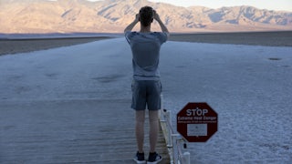 A shot of a man from the back, standing on a wooden platform taking a photo of the desert and mountains behind it. Next to him, a sign says STOP: EXTREME HEAT DANGER.