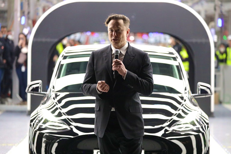 Elon Musk stands in front of a Tesla while speaking into a mic