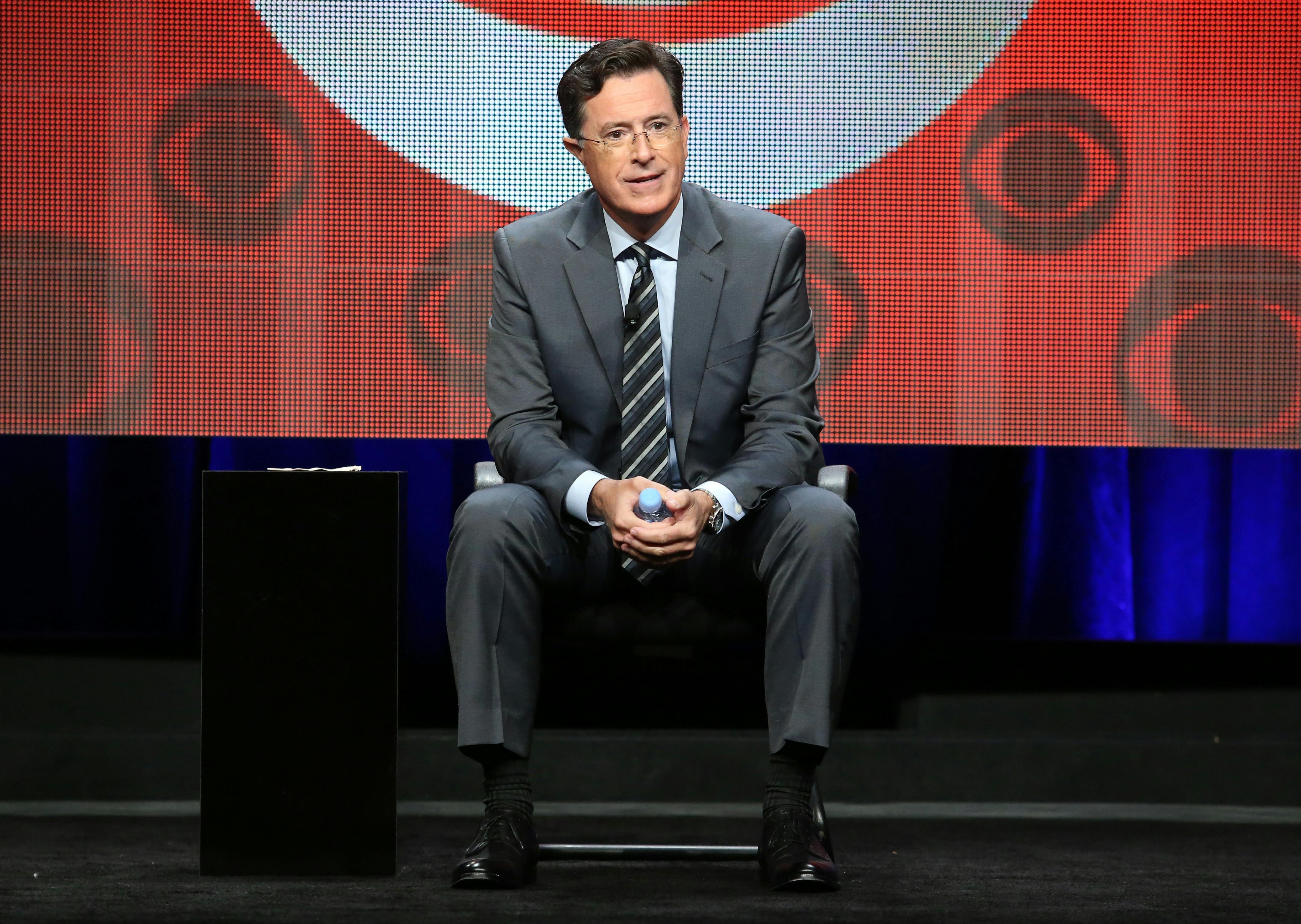 Stephen Colbert sits in front of a screen featuring several CBS logos