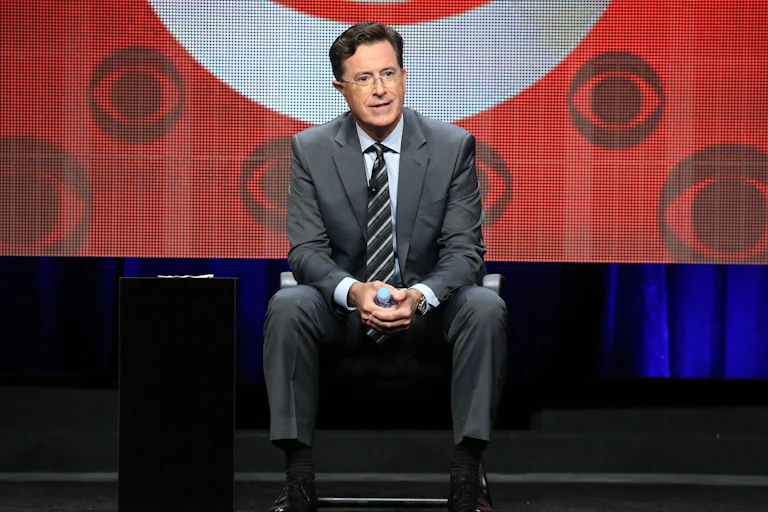 Stephen Colbert sits in front of a screen featuring several CBS logos