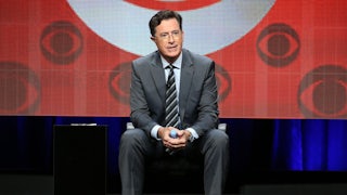 Stephen Colbert sits in front of a screen featuring several CBS logos