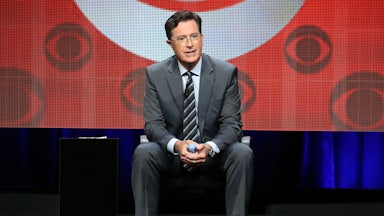 Stephen Colbert sits in front of a screen featuring several CBS logos