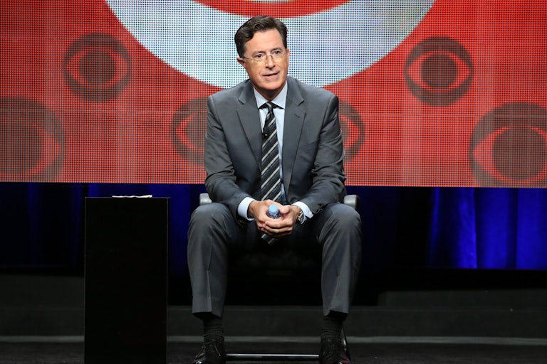 Stephen Colbert sits in front of a screen featuring several CBS logos