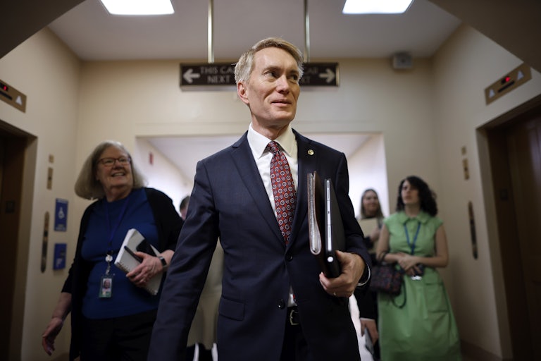 Senator James Lankford walks in the Capitol and smiles, binders in hand. Three women are in the background.