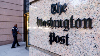 A marble pillar showing The Washington Post logo, with a security guard walking towards a revolving door behind it.