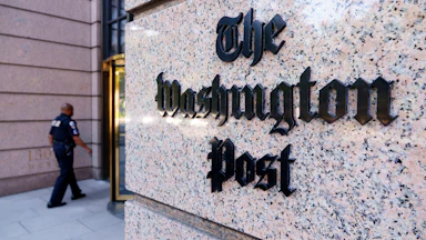 A marble pillar showing The Washington Post logo, with a security guard walking towards a revolving door behind it.