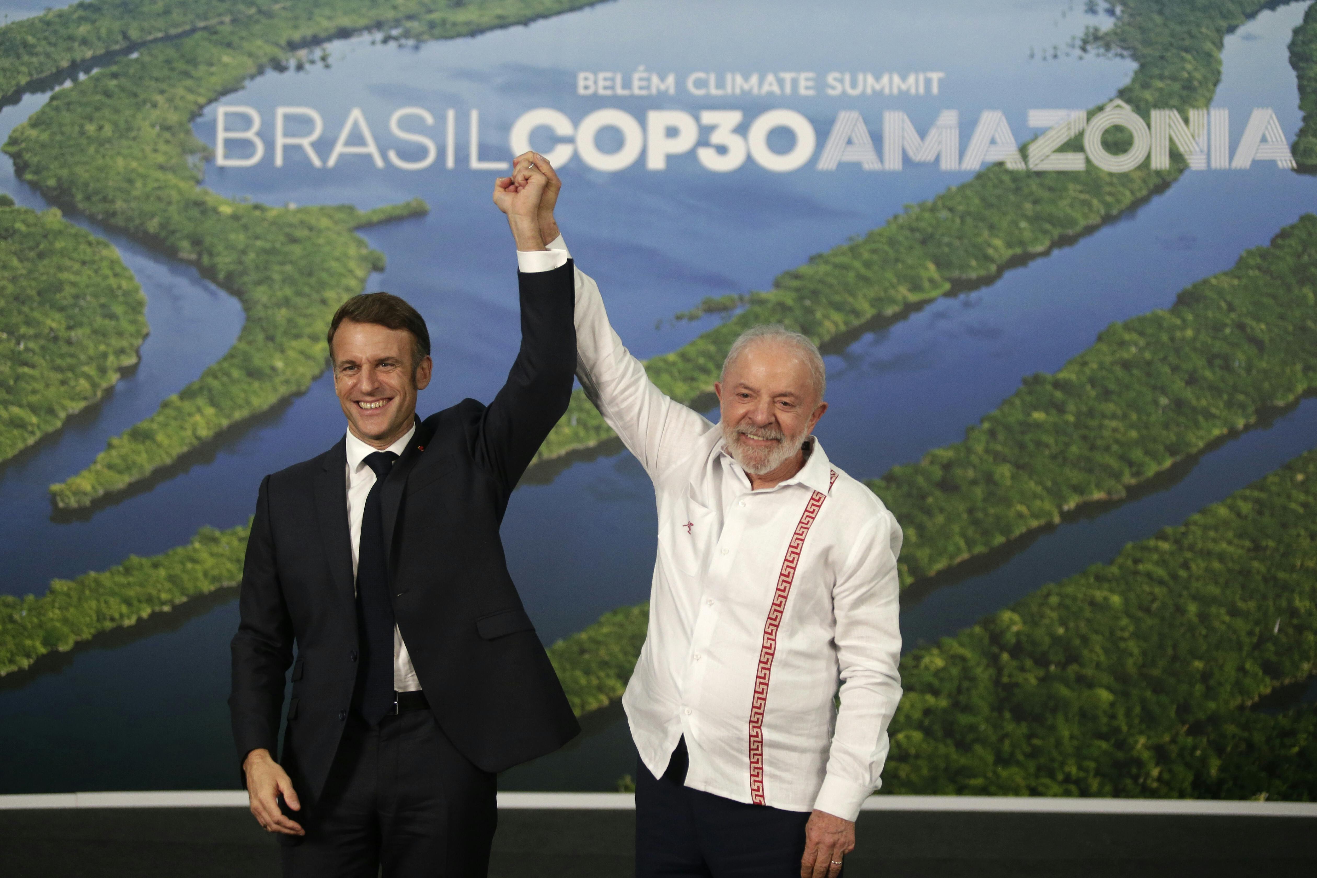 Lula and Macron stand holding their joined hands above their heads in front of a picture of the Amazon river.
