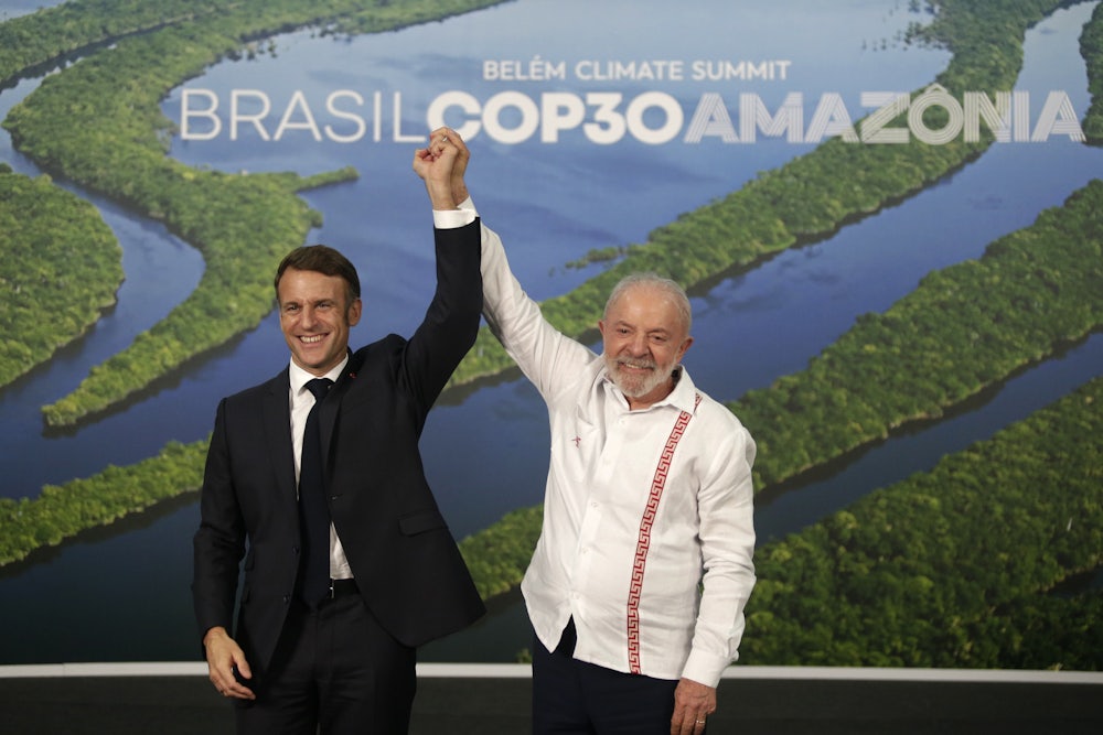 Lula and Macron stand holding their joined hands above their heads in front of a picture of the Amazon river.