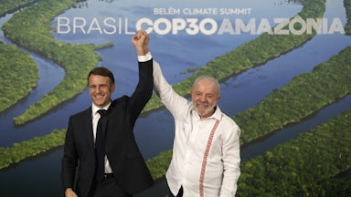 Lula and Macron stand holding their joined hands above their heads in front of a picture of the Amazon river.