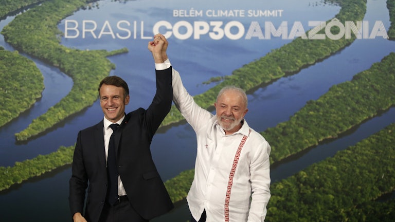 Lula and Macron stand holding their joined hands above their heads in front of a picture of the Amazon river.